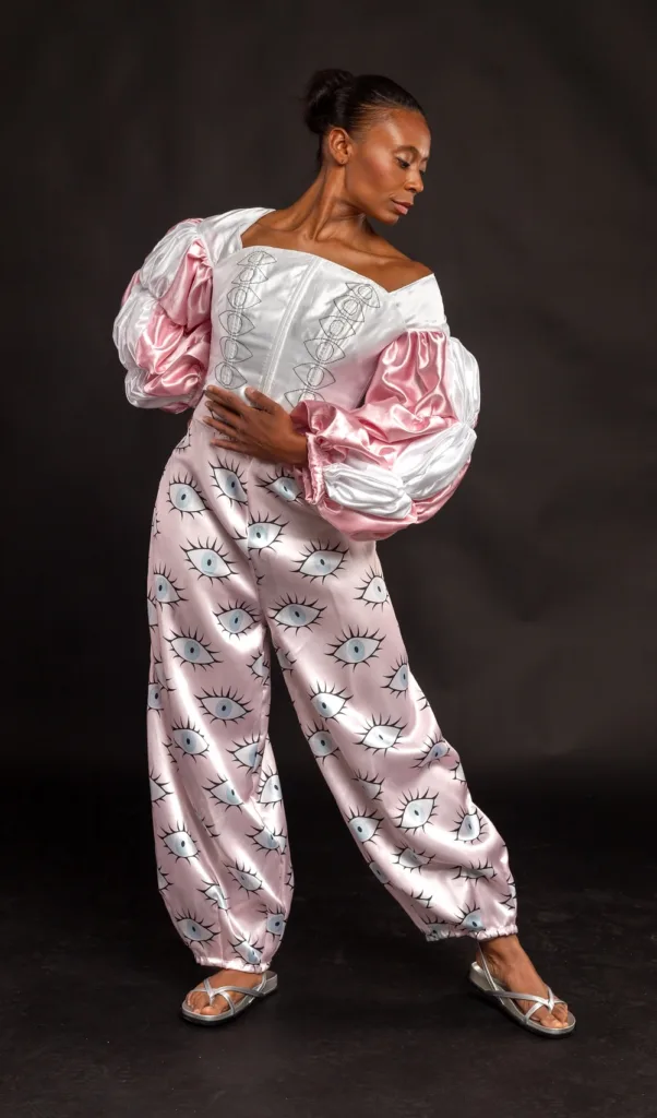 A fashion model stands looking down to the right in a full-length pose, showcasing a white satin corset-style top with pink puffy sleeves, loose pink satin pants printed with large eye motifs, and metallic silver flat sandals.