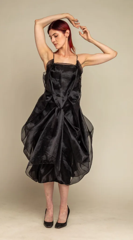Model in a sculptural black organza dress with a gathered, billowing skirt, arms raised on a beige backdrop